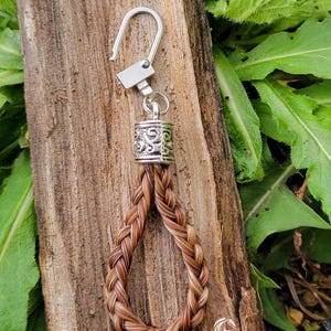 May include: A brown and white horsehair keychain with a silver charm and a silver hook. The keychain is braided and has a loop at the bottom. The keychain is on a wooden surface with green leaves in the background.  Equine Keepsakes horse hair jewelry.
