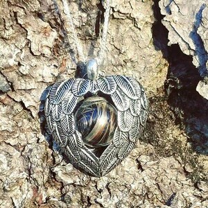 May include: A silver heart-shaped pendant with angel wings surrounding a clear glass orb filled with brown and black hair. The pendant is hanging from a silver chain against a rough bark background.