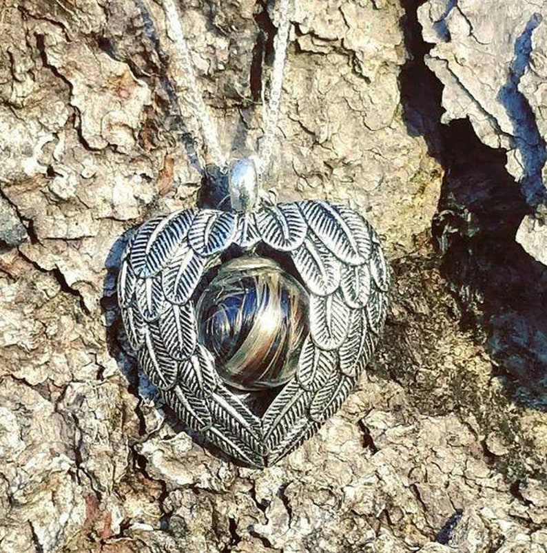 May include: A silver heart-shaped pendant with angel wings surrounding a clear glass orb filled with brown and black hair. The pendant is hanging from a silver chain against a rough bark background.