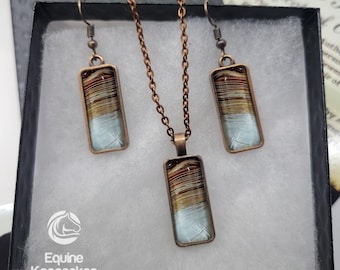 Puede incluir: Un conjunto de joyas de crin de caballo en una caja negra. El conjunto incluye un collar y pendientes. El collar y los pendientes tienen un colgante rectangular con un patrón marrón y blanco. El colgante está hecho de crin de caballo y resina. Las joyas están empaquetadas en una caja negra con el logotipo de la empresa y las palabras "Equine Keepsakes horse hair jewelry".
