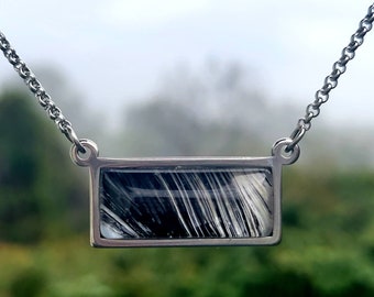 May include: A silver chain necklace with a rectangular pendant featuring a black and white abstract design.