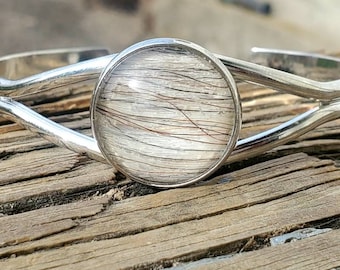 May include: A silver bracelet with a round, clear, glass-like charm containing a swirl of light brown and white hair.
