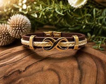 May include: A brown leather bracelet with a gold infinity symbol and accents. The bracelet features a braided cream-colored center section and gold-colored metal clasps. The bracelet is displayed on a wooden surface with greenery in the background.
