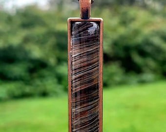 May include: A copper chain necklace with a rectangular pendant. The pendant is made of copper and has a clear glass insert with brown and white swirls.