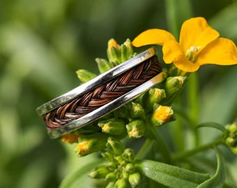 May include: A silver ring with a braided brown and copper-colored center band. The ring is set against a backdrop of green foliage and a bright yellow flower, creating a natural and elegant aesthetic.