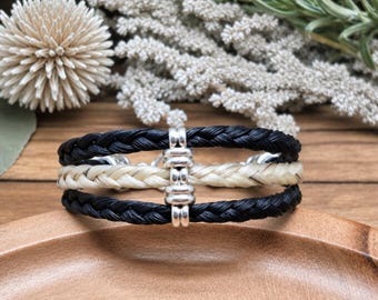 May include: A bracelet featuring three braided strands, with two outer black strands and one central cream-colored strand. The strands are held together by a silver-toned metal connector piece that secures the three braids in a horizontal alignment.