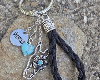 May include: A silver keyring with a braided black loop. Charms include a horse head, a turquoise heart, and a round tag with "Prosecco" engraved. The horse head has a flower with a turquoise centre. The keyring rests on a stone.