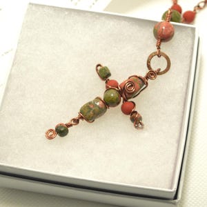 May include: A handmade copper wire cross pendant necklace with red and green stone beads. The cross is made of copper wire and features a unique design with wrapped wire and stone beads. The necklace is presented in a white gift box.