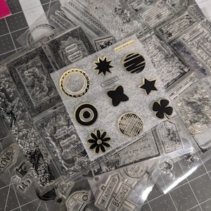 May include: Clear acrylic stamps with various designs, including floral, geometric, and travel-themed images. Some stamps feature text like "You are an AMAZING Woman" and "THE ADVENTURE BEGINS". The stamps are in clear plastic packaging.