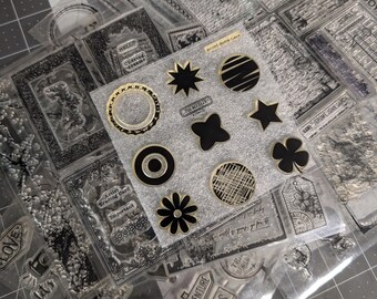 May include: Clear acrylic stamps with various designs, including floral, geometric, and travel-themed images. Some stamps feature text like "You are an AMAZING Woman" and "THE ADVENTURE BEGINS". The stamps are in clear plastic packaging.