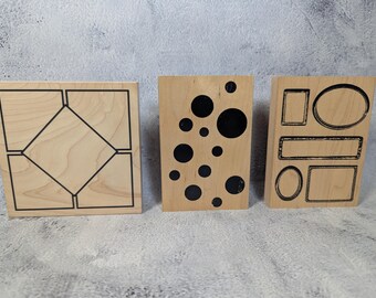 May include: Three wooden stamps with black designs. One stamp features a geometric pattern, another has black circles of varying sizes, and the third displays rectangular and oval frames. The stamps are on a light-colored surface.