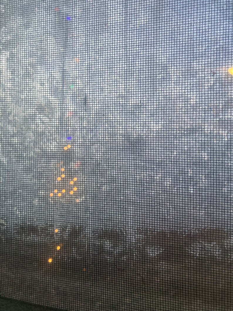 Pu&ograve; includere: Primo piano di una porta a zanzariera grigia con un motivo a griglia. La zanzariera &egrave; leggermente sporca e presenta alcuni piccoli fori. Attraverso la zanzariera sono visibili diverse piccole luci rotonde gialle.