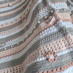 Könnte beinhalten: Eine gehäkelte Babydecke mit Streifen aus rosa, weißem und grauem Garn. Die Decke hat einen gewellten Rand und ist mit gehäkelten Blumen verziert.