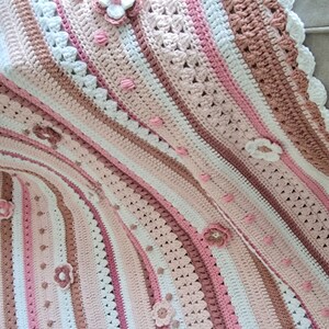 Könnte beinhalten: Eine gehäkelte Babydecke mit einem gestreiften Muster in Rosa, Weiß und Braun. Die Decke hat einen gewellten Rand und ist mit gehäkelten Blumen verziert.