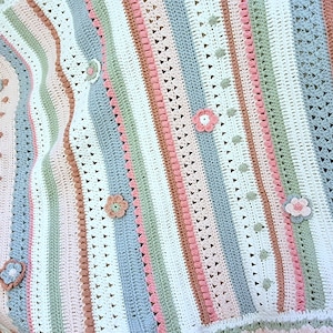 Könnte beinhalten: Handgefertigte Häkeldecke mit vertikalen Streifen in Pastelltönen von Weiß, Rosa, Blau und Grün. Die Decke ist mit gehäkelten Blumen und einem gewellten Rand verziert. Ein gemütliches Wohnaccessoire.