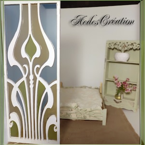 May include: A white, decorative room divider with a geometric design featuring green, brown, and blue glass panels. The divider is in front of a white wall and a miniature bed with a white bedspread.