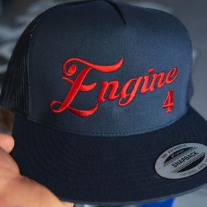 May include: A navy blue trucker hat with a red embroidered logo that says "Engine 4". The hat has a black mesh back and a black snap closure.