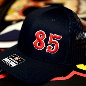 May include: A black and blue trucker hat with a red embroidered number "85" on the front. The hat has a mesh back and a curved brim.
