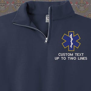 May include: A navy blue 1/4 zip sweatshirt with a white embroidered medical star of life symbol and the text "CUSTOM TEXT UP TO TWO LINES".
