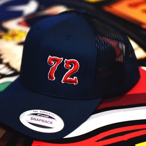 May include: A navy blue trucker hat with a red mesh back. The front of the hat features a large red embroidered number "72" in a stylized font. The hat has a white label that reads "The Authentic Snapback".