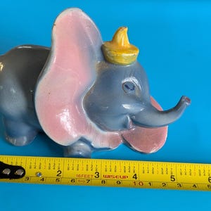May include: A ceramic Dumbo figurine, painted in gray, pink, and yellow. The elephant has large pink ears and a yellow hat. The figurine is approximately 4 inches long, as measured by a yellow tape measure.
