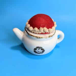 Puede incluir: Un cojín para alfileres con forma de tetera de cerámica blanca con una parte superior de fieltro rojo y un ribete decorativo. La tetera tiene un borde dorado y un diseño floral con el texto "LOVE YOU MOM".