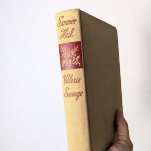 May include: A vintage book titled "Summer Hail" by Valerie Savage. The book has a tan cover with red lettering and a floral design. The spine displays the title and author's name in a serif font. The book is held in a hand.