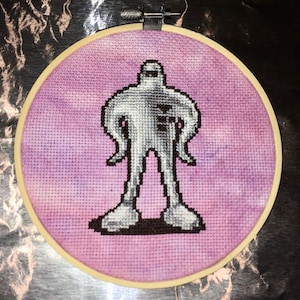 May include: A cross-stitch embroidery hoop with a purple background and a white figure with black outlines. The figure is standing with its arms at its sides.
