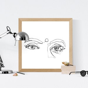 May include: A black and white line drawing of a woman's face with a focus on the eyes. The drawing is framed in a light wood frame and is displayed on a white surface.