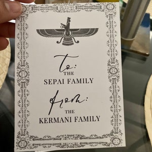 Pode incluir: Um cartão branco com uma imagem em preto e branco de um símbolo persa. O cartão tem uma borda preta com um padrão decorativo. O texto no cartão diz "To: The Sepai Family From: The Kermani Family".