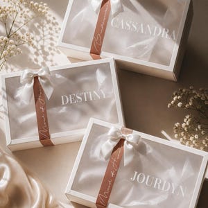 May include: White gift boxes with clear lids, each decorated with a white satin bow and a ribbon. The boxes are labelled with names: Cassandra, Destiny, and Jourdyn. The ribbon reads "Maid of Honour".