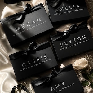 May include: Five black gift boxes, each with a name in white script and tied with a black satin ribbon. The boxes are on a satin surface, with the words "will you be my bridesmaid" below each name. A white rose and small white flowers are also present.