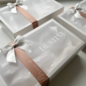 May include: Three white gift boxes with clear lids are arranged on a white surface. Each box is adorned with a white satin bow and a blush-colored ribbon. The names "Cassandre", "Jourdyn", and "Destiny" are printed on the boxes.