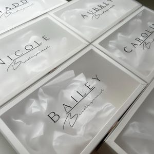 May include: Several white gift boxes with clear lids are arranged in a grid. Each box has a name printed in black script, with the word "Bridesmaid" below. The boxes are filled with white tissue paper.