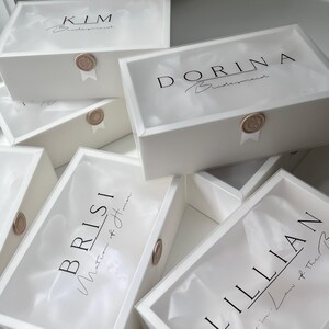 May include: White gift boxes with clear lids, each labeled with a name in black script. The boxes are sealed with a gold wax stamp and ribbon. The names include "KIM", "DORINA", "BRISI", and "LILLIAN".