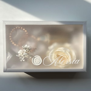May include: A clear rectangular box with a frosted front, containing a gold bracelet, white flowers, a wax seal, and a white rose. The name "Alessia" is written on the front of the box in a cursive font.