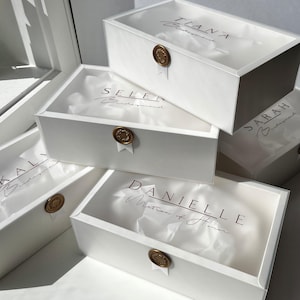 May include: Several white gift boxes with names printed on the lids. Each box is sealed with a gold wax seal and contains tissue paper. The names include Tiana, Selen, Sarah, Kal, and Danielle.