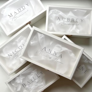 May include: White gift boxes with clear lids, each labeled with a name in black script. Some boxes include the word "Bridesmaid" or "Maid of Honor". The boxes are filled with white tissue paper.