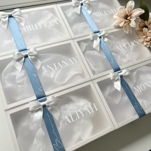 May include: White gift boxes with clear lids, each personalized with a name in white lettering. A light blue ribbon with white text and a white bow adorns each box. The boxes are arranged in a grid, suggesting a collection of gifts.