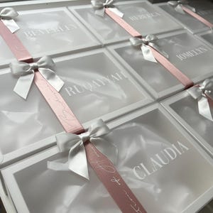 May include: White gift boxes with clear lids, each adorned with a white bow and a pink ribbon. The lids are printed with names in a white font. The boxes are arranged in a grid pattern.