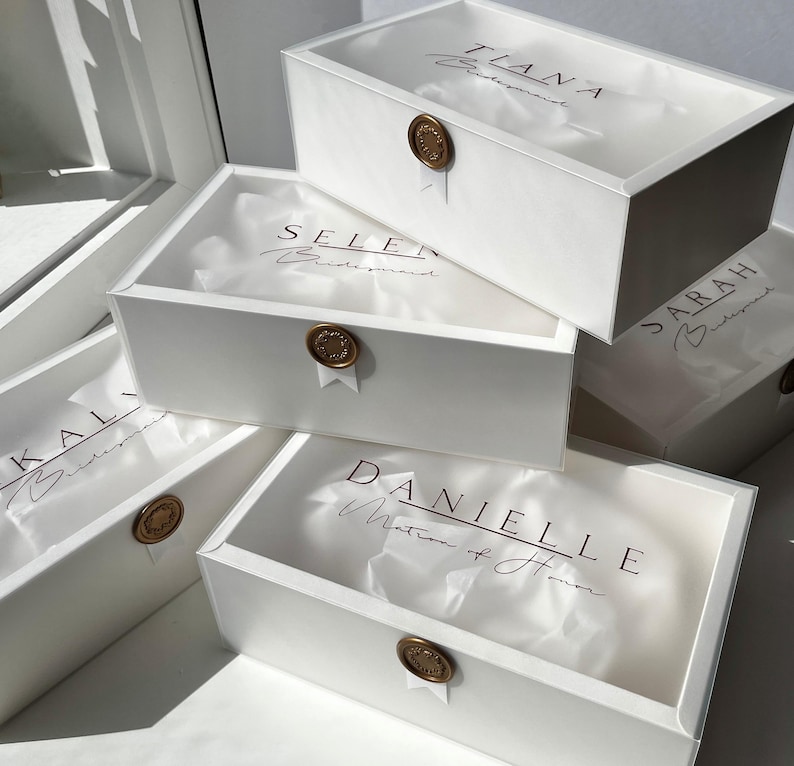 May include: Several white gift boxes with names printed on the lids. Each box is sealed with a gold wax seal and contains tissue paper. The names include Tiana, Selen, Sarah, Kal, and Danielle.
