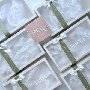 May include: White gift boxes with clear lids, each personalized with a name in white lettering. The boxes are tied with white satin bows and olive green ribbons. A pink square card with the text "ARIALA" is also present.