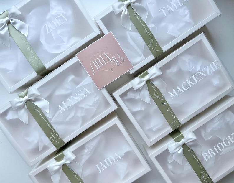 May include: White gift boxes with clear lids, each personalized with a name in white lettering. The boxes are tied with white satin bows and olive green ribbons. A pink square card with the text "ARIALA" is also present.