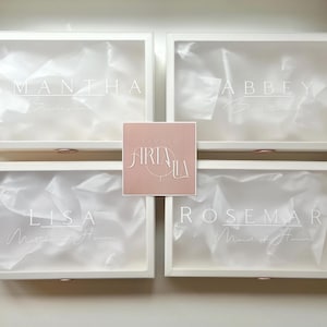 May include: Four white boxes with clear lids, each labeled with a name in elegant script: Samantha, Abbey, Lisa, and Rosemary. The boxes contain white tissue paper. A pink square with the logo "Airia Lila" is centered.