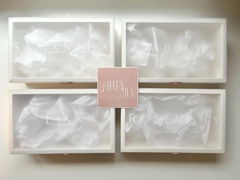 May include: Four white boxes with clear lids, each labeled with a name in elegant script: Samantha, Abbey, Lisa, and Rosemary. The boxes contain white tissue paper. A pink square with the logo "Airia Lila" is centered.