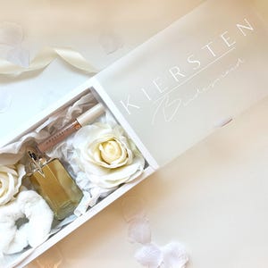 May include: A white gift box with the name "KIERSTEN" and the word "Bridesmaid" printed on the lid. Inside are white roses, a scrunchie, a bottle of perfume, and a tube of lip gloss. The box is on a white surface with scattered petals.