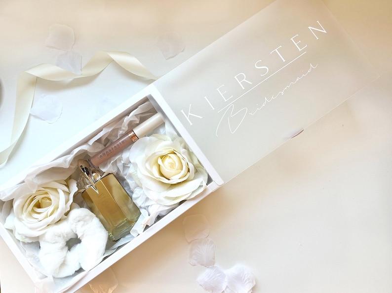 May include: A white gift box with the name "KIERSTEN" and the word "Bridesmaid" printed on the lid. Inside are white roses, a scrunchie, a bottle of perfume, and a tube of lip gloss. The box is on a white surface with scattered petals.