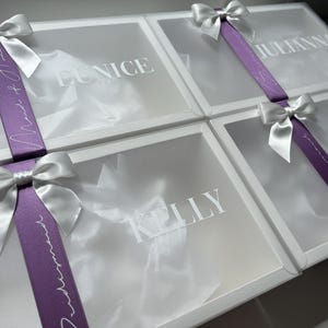 May include: Four white gift boxes with clear lids, each adorned with a white satin bow. A purple ribbon with white script wraps around each box. Names are printed on the boxes: Eunice, Julianna, Kelly, and Jan.