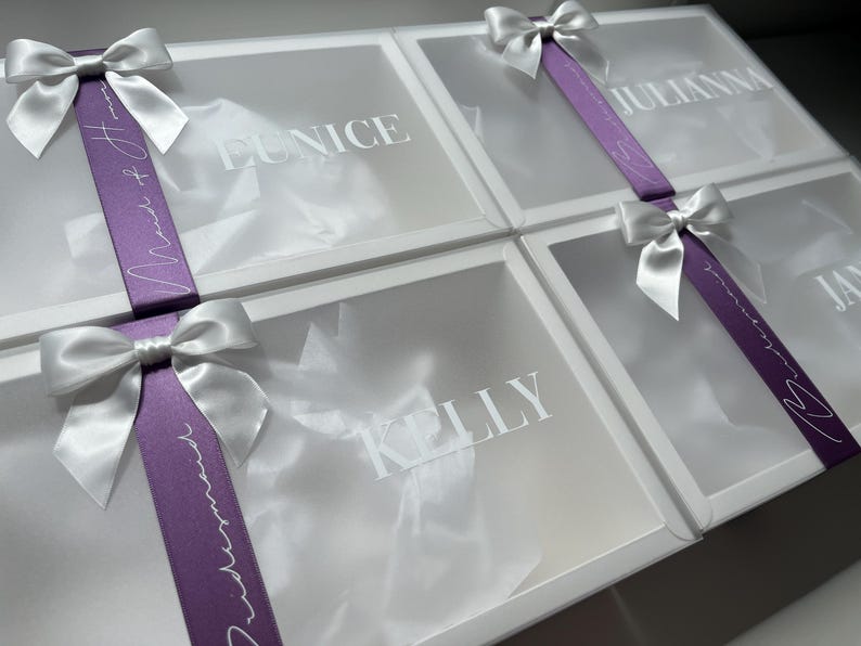 May include: Four white gift boxes with clear lids, each adorned with a white satin bow. A purple ribbon with white script wraps around each box. Names are printed on the boxes: Eunice, Julianna, Kelly, and Jan.