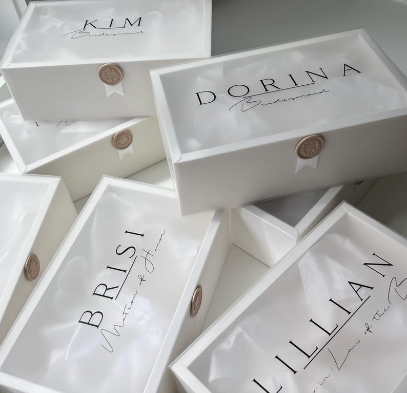 May include: White gift boxes with clear lids, each labeled with a name in black script. The boxes are sealed with a gold wax stamp and ribbon. The names include "KIM", "DORINA", "BRISI", and "LILLIAN".